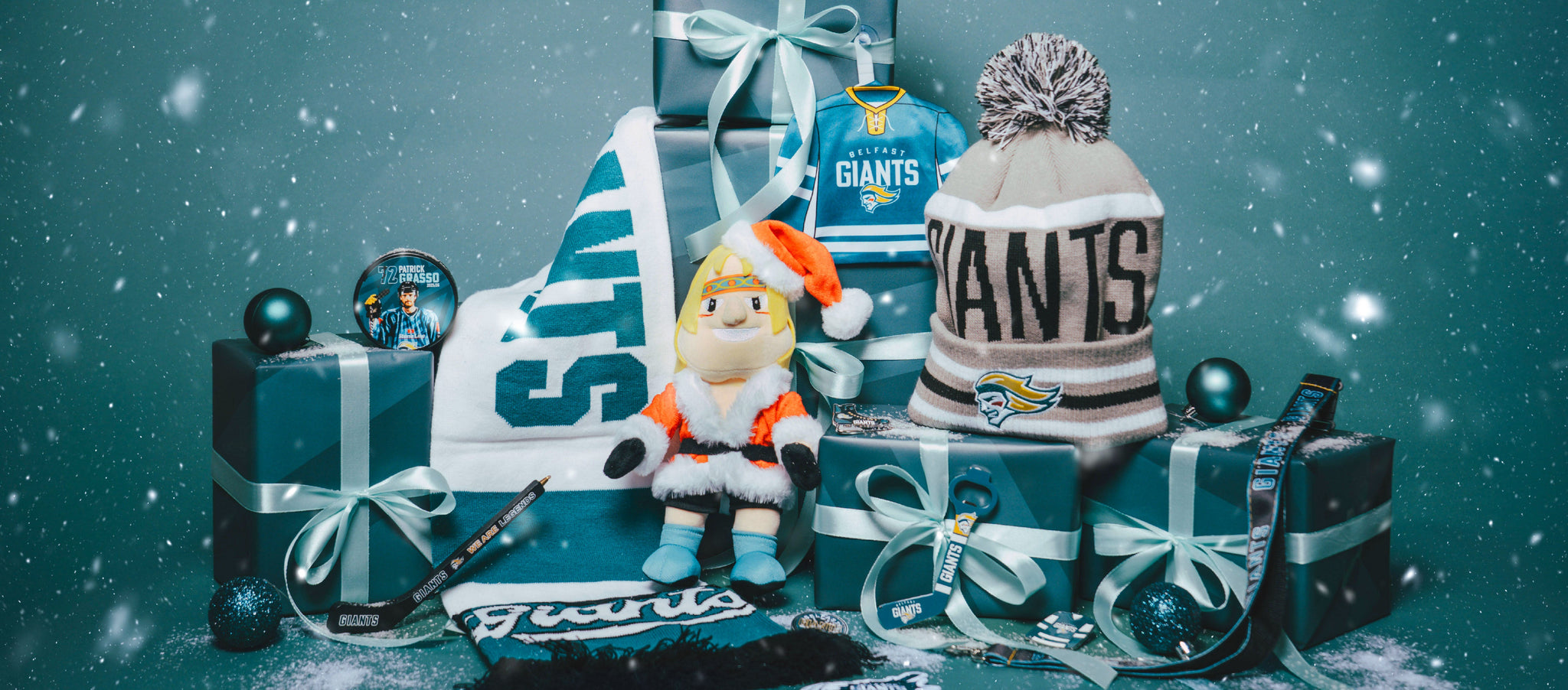 A festive arrangement of Cardiff Giants merchandise, including a plush mascot, scarf, beanie, ornaments, and gift-wrapped boxes, all dusted with snowflakes against a teal background.