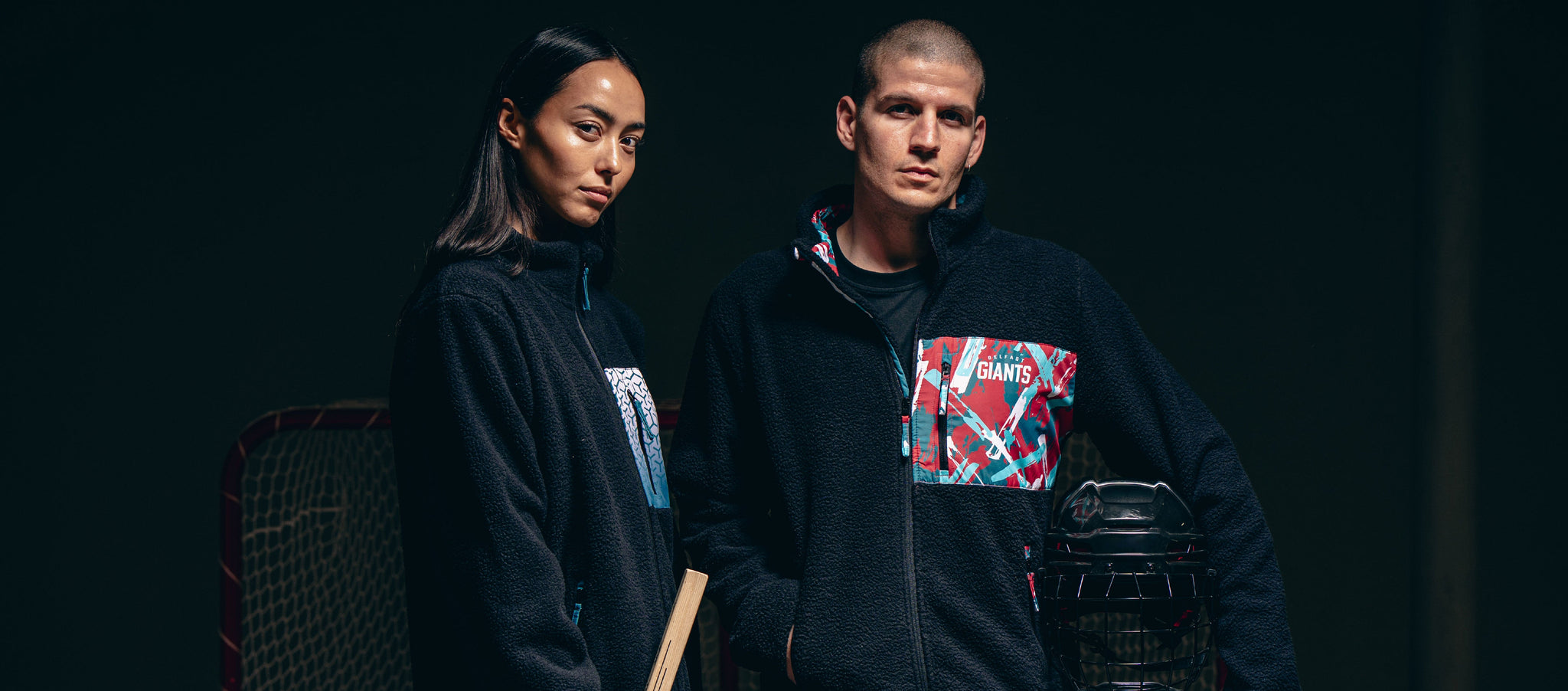 Two people wearing black zip-up jackets with colorful patterned pockets stand side by side against a dark background, with sports equipment partially visible behind them.
