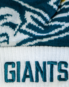 Close-up of the Belfast Giants Hockey Beanie Off-White by Belfast Giants, featuring "GIANTS" in teal on a cosy white knit and a stylized face pattern—ideal for winter matchdays.