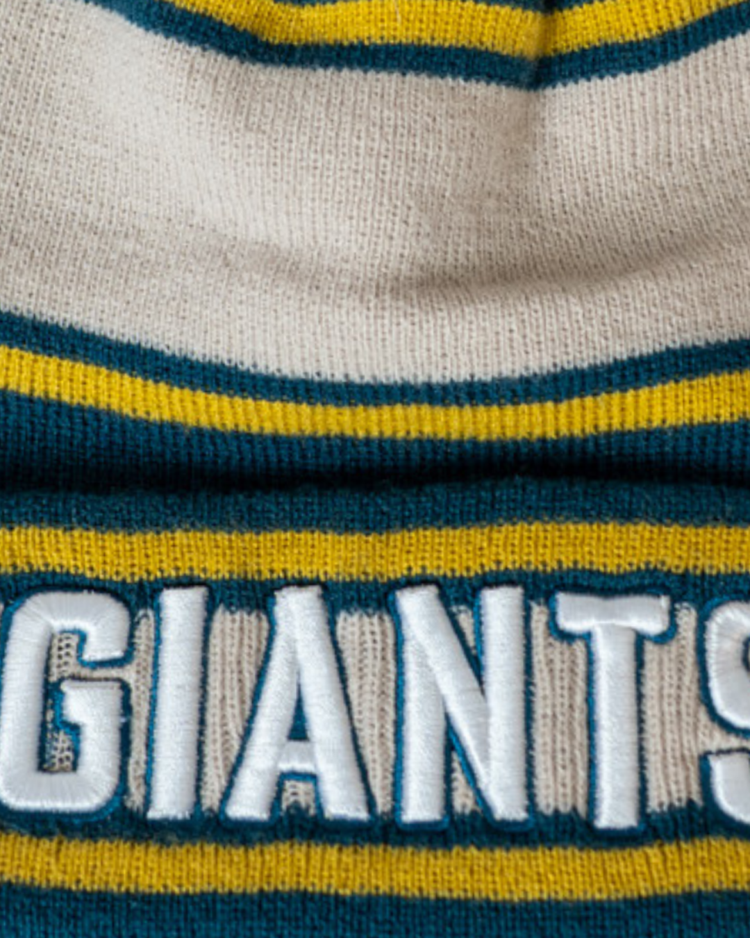 Close-up of the Belfast Giants Beanie Cream/Teal by Belfast Giants, a knit hat with cream, teal, and yellow stripes, featuring "GIANTS" embroidered in large white letters.