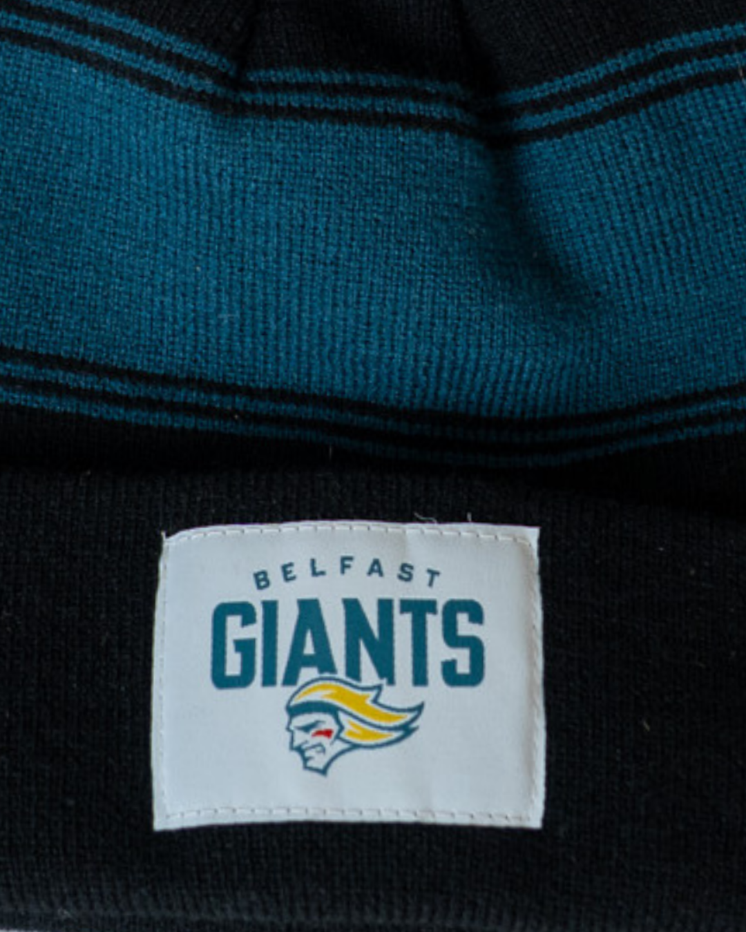 A close-up of the Belfast Giants Black Beanie Teal Stripes, featuring teal and black stripes and a stitched Belfast Giants patch above the team's bold giant face logo.