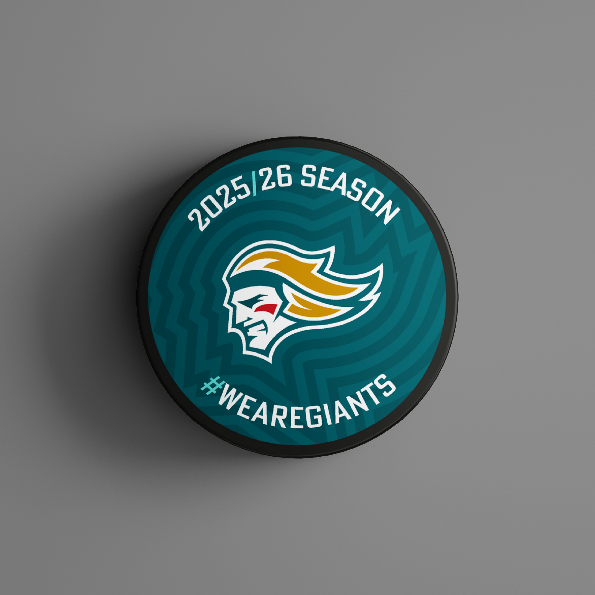The Logo Season Puck Gold 25-26 from Belfast Giants Store by National Merchandise features a warrior head logo, “2025/26 Season” at the top, and “#WEAREGIANTS” at the bottom on a gray and teal patterned background.
