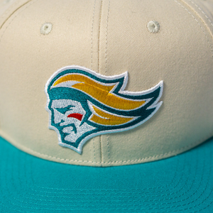 The Belfast Giants Hockey Cap Off-White is an adjustable beige and teal cap featuring the Belfast Giants embroidered logo—a stylized face with gold hair, teal outline, and a red cheek accent.