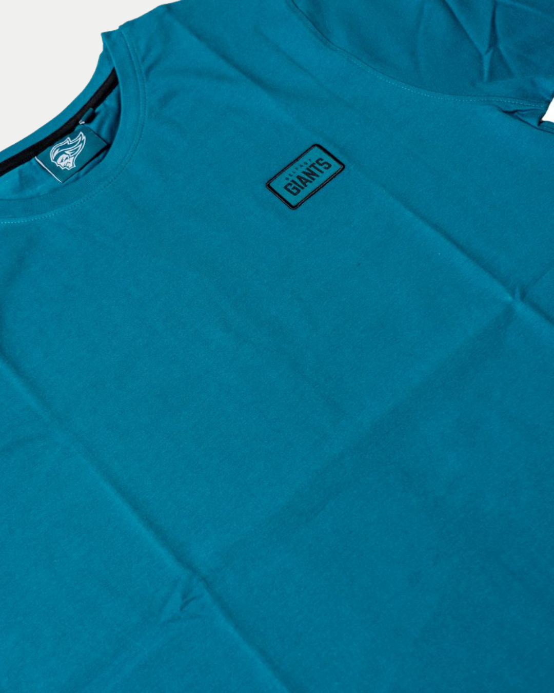 The Belfast Giants Teal T-Shirt Black Badge features a crew neck, soft teal fabric with a subtle wrinkled look, and a small rectangular chest patch displaying "GIANTS" in bold black letters.