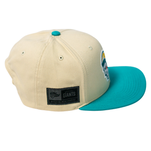 The Belfast Giants Hockey Cap Off-White features a teal brim and button, a Giants Official Merchandise patch, and a colorful side logo. This adjustable Belfast Giants hat is shown on a transparent background.