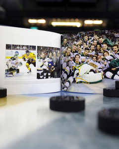 Ring the Bell! The Story of the Friendship Four by Belfast Giants is open on a hockey rink, showing NCAA Division One team moments. Black hockey pucks are scattered around the book on the ice.
