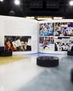 A copy of "Ring the Bell! The Story of the Friendship Four" by Belfast Giants is open on an ice rink with hockey pucks in front, displaying images of cheering fans and supportive signs at a NCAA Division One college hockey tournament.