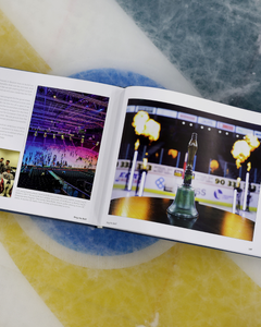 An open copy of "Ring the Bell! The Story of the Friendship Four" by Belfast Giants sits on an ice hockey rink, its pages displaying vibrant photos—one of a flaming trophy, another of a lit NCAA Division One tournament arena.