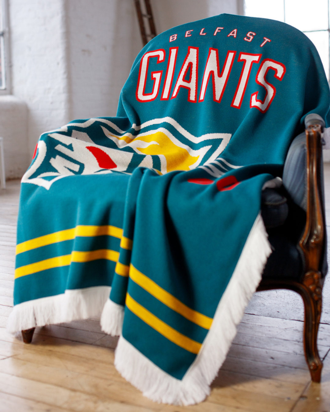 The Belfast Giants Blanket, by Belfast Giants, is a blue recycled acrylic throw featuring the team’s logo, bold red and white lettering, and yellow stripes, draped over a dark armchair in a sunlit room with wooden floors.